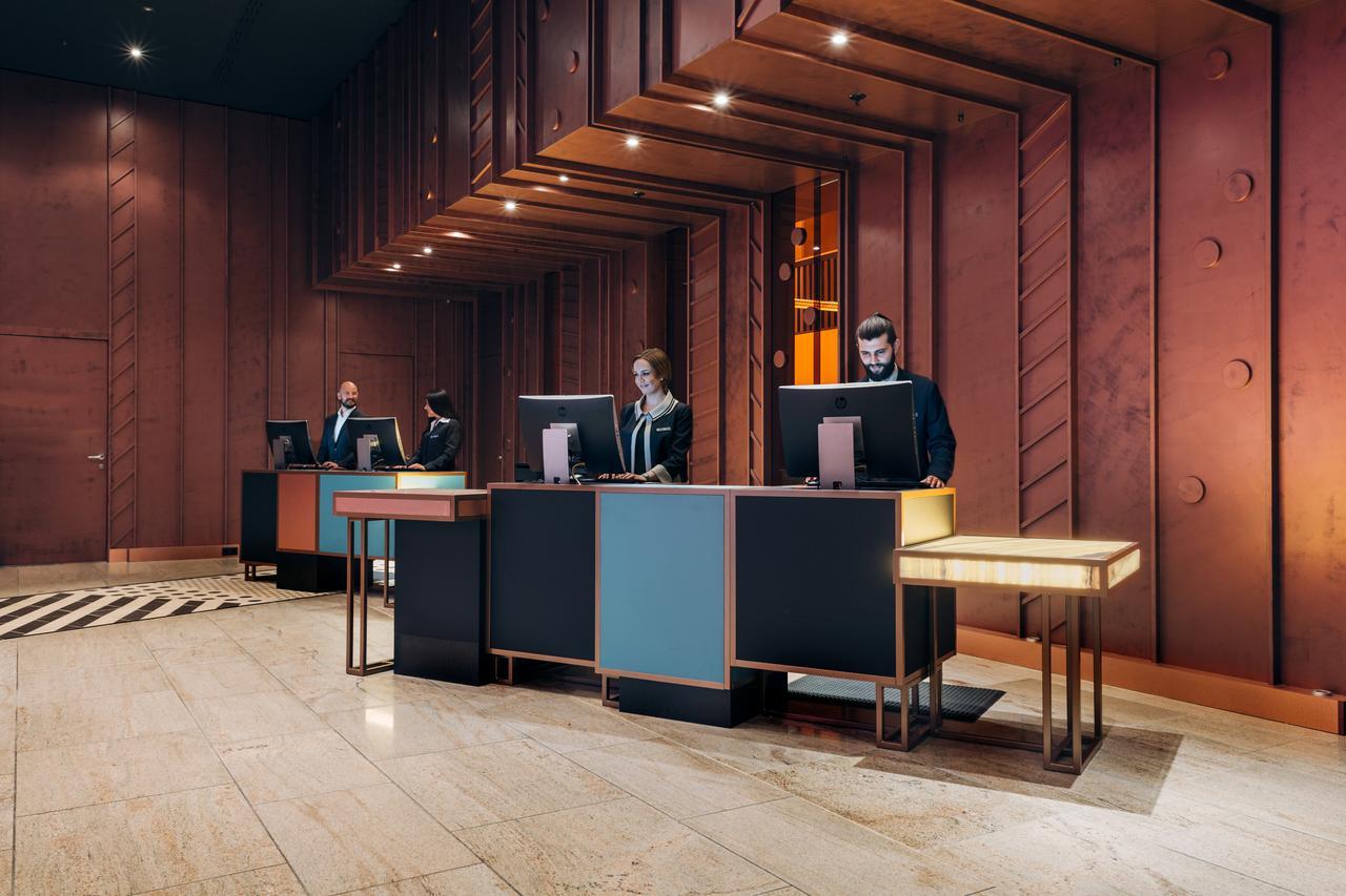 Pullman Berlin Schweizerhof Hotel Bagian luar foto The image shows a modern hotel lobby reception area. There are several reception desks made of dark materials with illuminated accents. Behind the desks, three people are standing, likely staff members, engaged with computer screens. The lobby featur